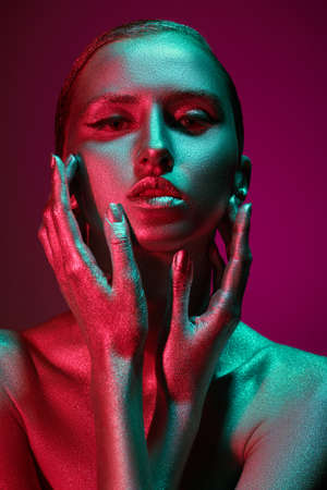 Beauty industry, cosmetology. Art portrait of a beautiful woman with shiny silver skin in colorful green and red light on a fuchsia background. Beauty and cosmetics. Body painting.の写真素材