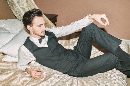 A handsome glamorous young man in a stylish three-piece suit and a bow tie is lying relaxed on a bed...