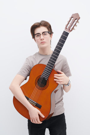 A handsome teenage boy in glasses and a grey T-shirt poses on a white background holding an acoustic...
