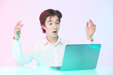 A puzzled teen boy in a neat white shirt looks with surprise at the screen of his laptop and spreads...