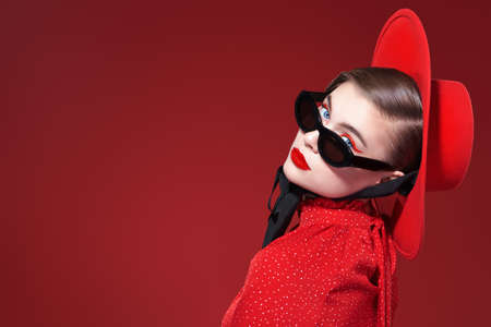 A stylish pretty girl model with bright makeup poses in feminine red dress, red hat and stylish sunglasses. Red background. Fashion and beauty.の写真素材