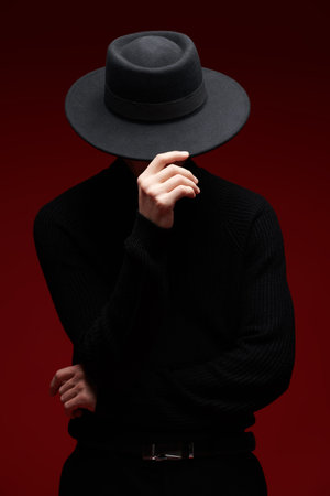 Young brunet man in a black turtleneck and a hat hiding his face poses on a red studio background....