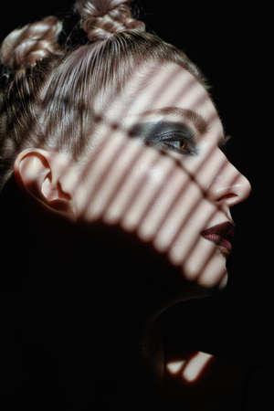 Make up and cosmetics. Close-up portrait of a fashion model girl with dark makeup posing in shadows. Studio shot on a black background. Sideview shot.の写真素材