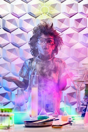Education, science and children. A funny excited little boy with glasses and matted hair stands in a chemical laboratory after explosion with surprise on his face.の写真素材