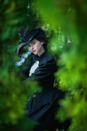 Portrait of a charming lady in a black dress and hat walking among green foliage in the park. Historical makeup and hairstyle. Historical reconstruction of the late 19th and early 20th centuries.の写真素材