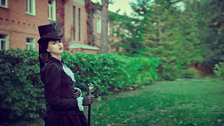 A confident and sophisticated lady in a strict black suit of the 19th century walks along a brick house surrounded by green foliage. Copy space.の写真素材