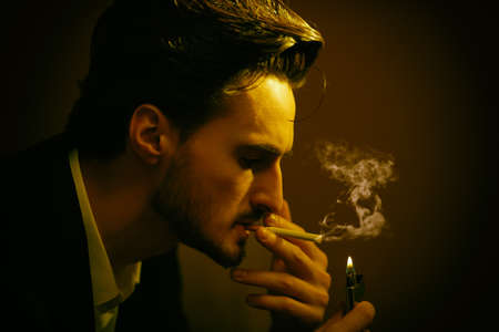 A confident handsome brunet man in an elegant black suit lights a cigarette in a dark room. Noir style. Male beauty, fashion.の写真素材