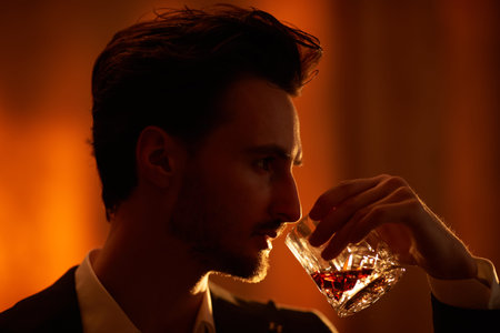 Sideview shot of handsome brunet man standing in a room with dark lighting and drinking whiskey....