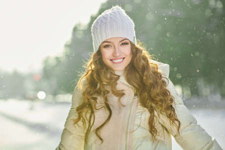 Pretty girl in a warm winter down jacket and a knitted hat rejoices on a sunny winter day with snowfall. Winter fashion. Good mood, emotions.の写真素材