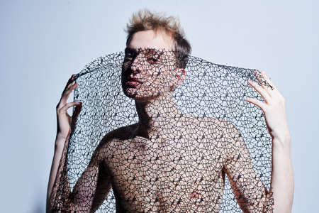 Art and fashion. Young man fashion model with naked torso poses using black mesh fabric in the studio on a grey background.の写真素材