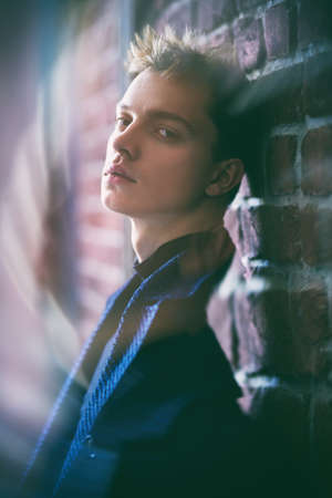Life, people and emotions. Portrait of a young man in a coat, who stands leaning against a brick wall and looks sadly at the camera. Blur effects.の写真素材