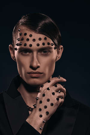 Fashion concept. Portrait of a handsome brutal man in a black tailcoat with metal spikes on his face and arm on a black background.の写真素材