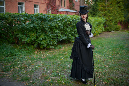 A pretty, sophisticated lady in a strict black suit of the 19th century walks along a brick house surrounded by green foliage. Copy space.の写真素材