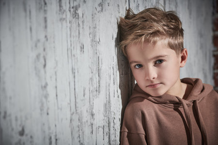Portrait of a cute blond boy in hoody posing in the studio leaning against a concrete wall. Modern...