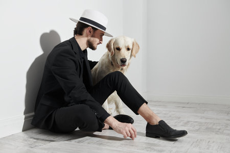 A handsome elegant brunette man in a black suit and white hat sits on the floor against a white wall...