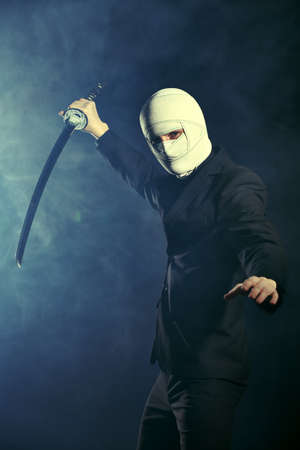 Business concept, Competitive wars.  A warrior businessman in a classic black suit, with a bandaged face, stands on alert with a katana samurai sword. Studio portrait on a dark background with smoke.の写真素材