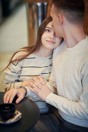 Pretty girl sits close to her boyfriend at a table in a cozy cafe holding hands and looking into...