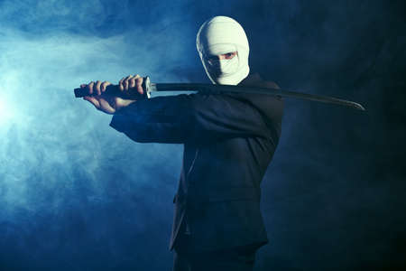 A warrior businessman in black suit, with a bandaged face, stands in combat readiness with a katana samurai sword. Competitive wars, business concept. Studio portrait on a dark background with smoke.の写真素材