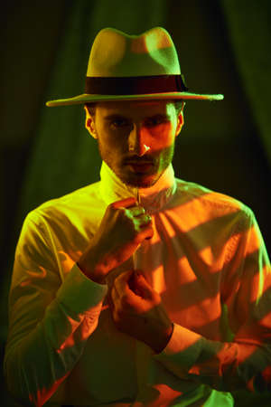 An elegantly dressed brunet man in white hat buttons his shirt standing in a dark bedroom. Mafia and the criminal world.の写真素材