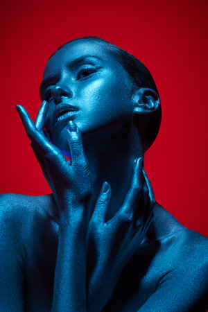 Portrait of a beautiful woman with painted shiny silvery skin posing with graceful hands. Studio shot on a red background in neon blue light. body painting. Beauty industry, cosmetology, cosmetics.の写真素材
