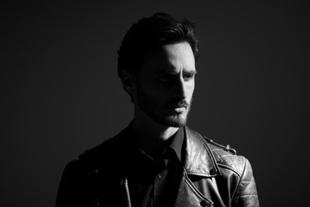 Black-and-white portrait of a brutal brunet man in a stylish leather jacket standing on a dark background frowning and looking intensely away. Male beauty, fashion. emotions.の写真素材