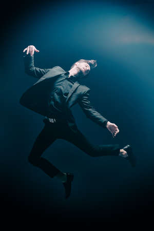 A man in a black classic suit jumps, feeling freedom and lightness. Studio full-length portrait on a dark blue background. Happiness. The concept of success and aspirations, dreams.の写真素材