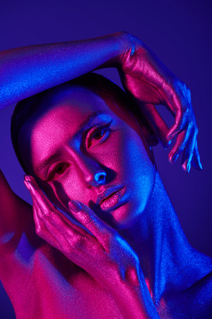 Studio shot of a beautiful fashion model with shiny silver skin posing gracefully in colorful neon blue and pink light. beauty and cosmetics concept. body painting. blue background.の写真素材