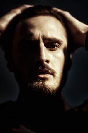 A handsome brutal brunet man in black clothes stands with his hands on his head on a dark background...