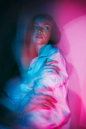 Portrait of a young beautiful girl with long hair in white shirt hugging herself and looking away. Blurred studio portrait in blue and crimson lighting.の写真素材