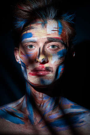 Art and male beauty. Portrait of a handsome man with colorful painted face standing on a black background with shadows.の写真素材