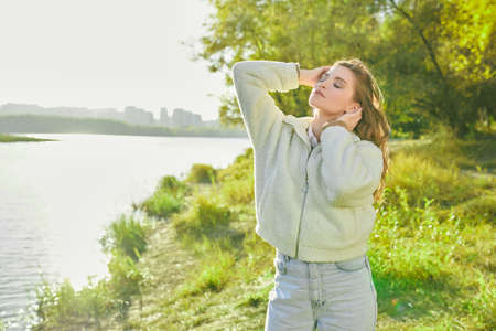 Pretty fair-haired girl in stylish casual clothes stands relaxed on the back of the river on a warm sunny day with closed eyes.の写真素材