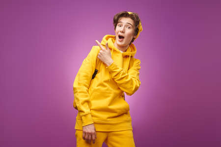 A cool modern teenager dressed in stylish bright youth clothes smiles and excitedly points his index finger away. Purple background with copy space. Youth lifestyle and emotions.の写真素材