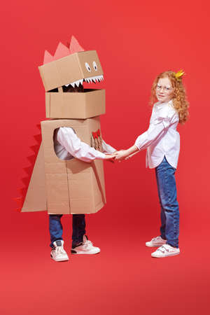 Children's fantasies and dreams. Two cute children in white shirts and jeans hold their hands playing cardboard dragon and princess. Full-length portrait on a red background.の写真素材