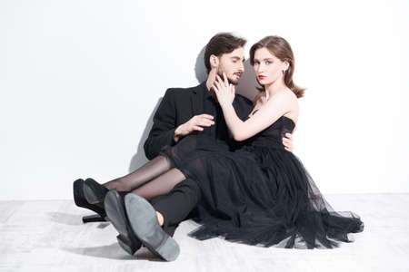 Full length portrait of a beautiful couple of lovers in black evening dresses sitting in each other's arms on the floor against a white wall. Evening fashion and beauty. Love relationship.の写真素材