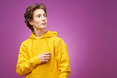 Portrait of a modern teenage boy in bright yellow hoodie looking away with interest. Purple background with copy space. Adolescents and lifestyle. Youth fashion.の写真素材