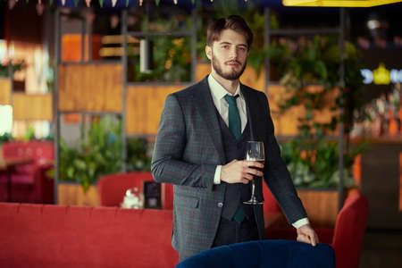 A confident handsome brunet man in elegant classic suit stands in a restaurant with a glass of wine. Men's beauty, fashion. Business style. Businessman.の写真素材