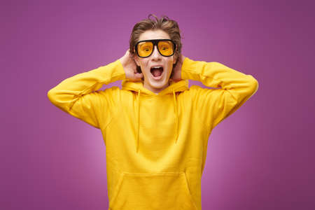 A handsome emotional teenager in bright youth clothes and stylish sunglasses screams holding his head with his hands. Purple background. Youth lifestyle and emotions.の写真素材