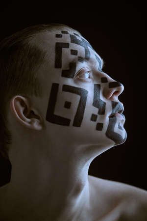 Sideview shot of a young man with a QR-code painted on his face looking away with a lost look. Studio portrait on a black background. Chipping and control over society concept.の写真素材