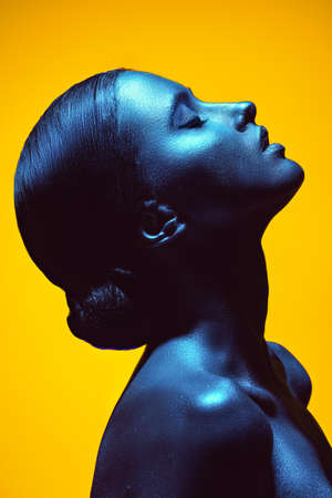 A beautiful woman with painted shiny silvery skin posing in profile with closed eyes. Studio shot on a yellow background in blue light. Body painting. Beauty industry, cosmetology, cosmetics.の写真素材