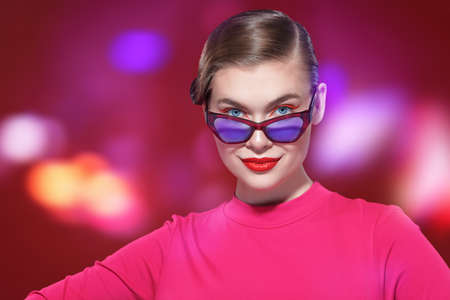 Beautiful elegant blonde girl with bright makeup and stylish glasses poses on a red background with colorful lights. Make up and cosmetics. Glasses style.の写真素材