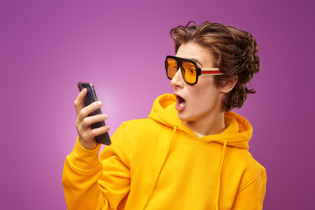 A surprised teenage boy in stylish sunglasses and yellow hoodie emotionally reacts to the received...