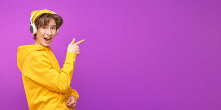 A cool modern teenager in stylish bright youth clothes and headphones smiles and joyfully points his index finger away. Purple background with copy space. Youth lifestyle and emotions.の写真素材