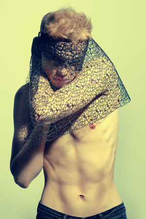 Art and fashion. Young man fashion model with naked torso holds black mesh fabric hiding his face....