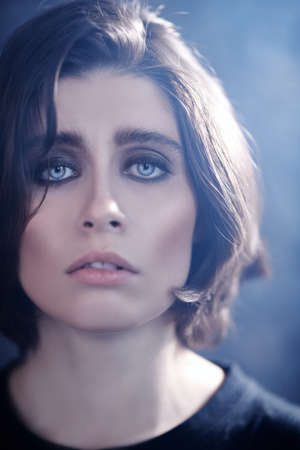 Portrait of a brunette girl with beautiful blue eyes and dramatic dark eye makeup looking sadly at the camera. Psychological picture. People, emotions.の写真素材