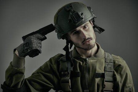 Portrait of a soldier in Protective Combat Uniform putting a gun to his head. Studio portrait on a...