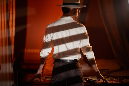 A masculine brunet man in a classic white shirt and hat stands with his back to the camera in a dark...