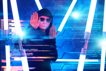 Teen boy hacker. A modern teen boy in a black gown, mask and black sunglasses hiding his face works with hologram in a dark space among neon lights. Futuristic technologies.の写真素材