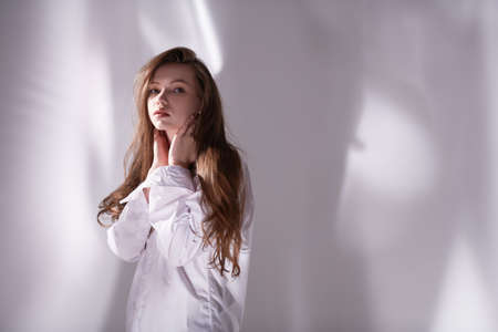 A young beautiful girl with long brown hair in a white shirt stands calmly and pensively on a white background with shadows. Copy space.の写真素材