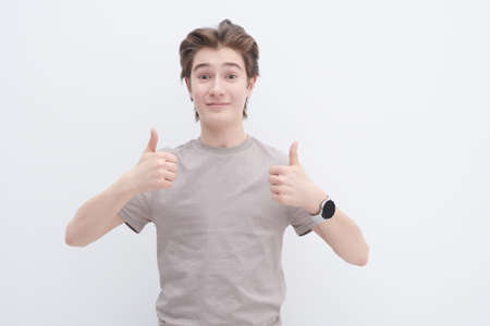 A handsome smiling teenage boy in a grey T-shirt joyfully shows his thumbs up. White background. Adolescents, emotions, lifestyle. Happy teenage boy.の写真素材
