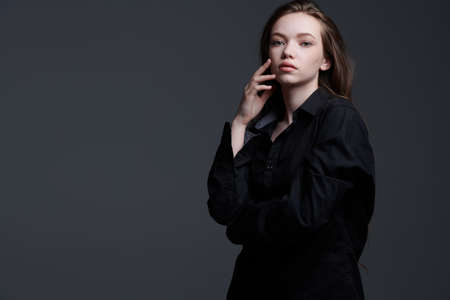 Beauty, people, emotions. Portrait of a beautiful young girl with long brown hair and in a black shirt, who looks calmly at the camera. Dark grey background with copy space.の写真素材